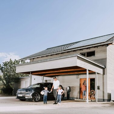 Familie steht vor Einfamilienhaus mit Photovoltaikanlage, Wärmepumpe und Wallbox
