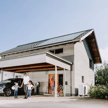 Familie steht vor Einfamilienhaus mit Photovoltaikanlage, Wärmepumpe und Wallbox