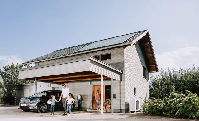 Familie steht vor Einfamilienhaus mit Photovoltaikanlage, Wärmepumpe und Wallbox