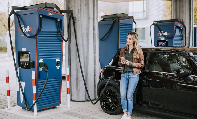 Frau lehnt sich an ihrem Auto an. während das Fahrzeug am Schnelllader auflädt