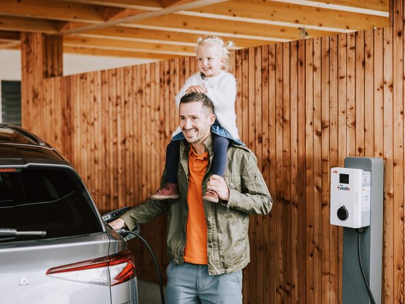 Mädchen sitzt auf den Schultern ihres Vaters, der sein E-Auto auflädt