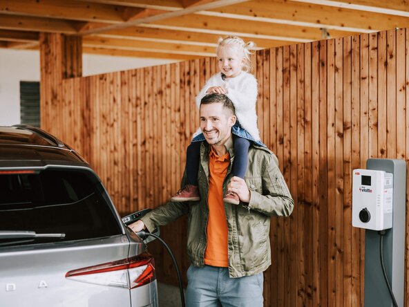 Mädchen sitzt auf den Schultern ihres Vaters, der sein E-Auto auflädt