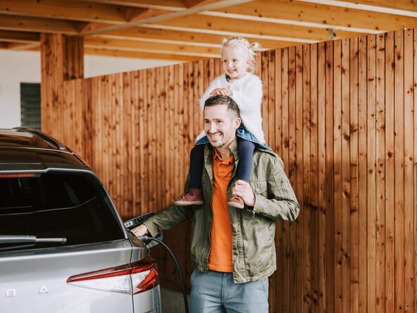 Mädchen sitzt auf den Schultern ihres Vaters, der sein E-Auto auflädt