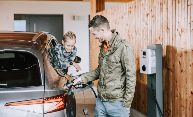 Mann lädt sein E-Auto an der Wallbox zuhause, während sein Sohn aus dem Autofenster sieht