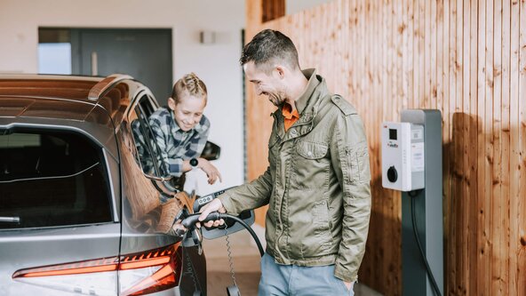 Mann lädt sein E-Auto an der Wallbox zuhause, während sein Sohn aus dem Autofenster sieht