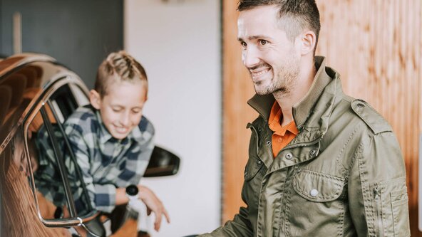 Mann lädt sein E-Auto an der Wallbox zuhause, während sein Sohn aus dem Autofenster sieht