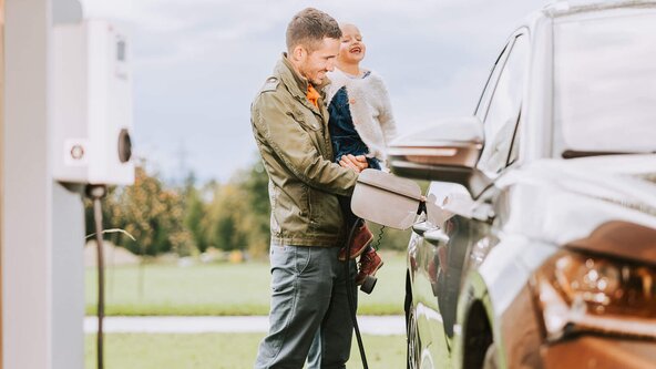 Vater mit Tochter im Arm lädt sein E-Auto an einer Wallbox auf