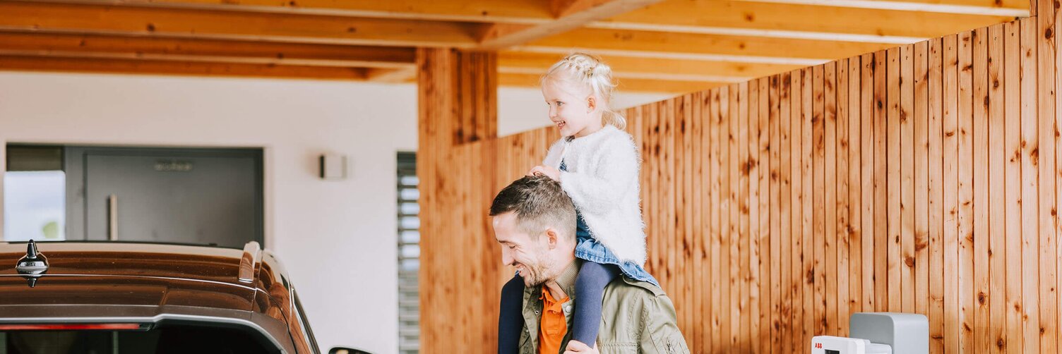 Mädchen sitzt auf den Schultern ihres Vaters, der sein E-Auto auflädt