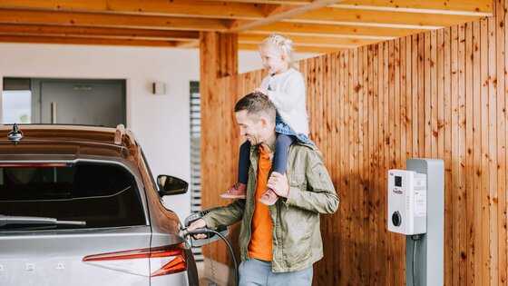 Mädchen sitzt auf den Schultern ihres Vaters, der sein E-Auto auflädt