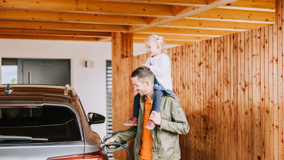 Mädchen sitzt auf den Schultern ihres Vaters, der sein E-Auto auflädt