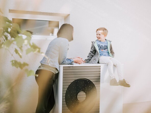 Ein Junge sitzt auf einer Wärmepumpe, die Mutter steht daneben